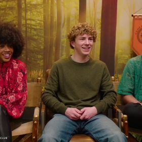 In an image from D23's Camp Half-Blood Superlatives with the Cast of Percy Jackson and the Olympians video, from left to right, Leah Sava Jeffries (Annabeth), Walker Scobell (Percy), and Aryan Simhadri (Grover) sit in director's chairs and smile either at someone off camera or at each other. Jeffries is wearing a red flowery blouse and leather pants; Scobell is in an army green sweater and jeans; and Simhadri is wearing a patterned green button-up shirt with black pants. Behind them is a backdrop reminiscent of campgrounds, and to Jeffries' left is both a shield and a sign with the show's logo on it.
