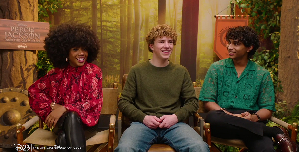 In an image from D23's Camp Half-Blood Superlatives with the Cast of Percy Jackson and the Olympians video, from left to right, Leah Sava Jeffries (Annabeth), Walker Scobell (Percy), and Aryan Simhadri (Grover) sit in director's chairs and smile either at someone off camera or at each other. Jeffries is wearing a red flowery blouse and leather pants; Scobell is in an army green sweater and jeans; and Simhadri is wearing a patterned green button-up shirt with black pants. Behind them is a backdrop reminiscent of campgrounds, and to Jeffries' left is both a shield and a sign with the show's logo on it.