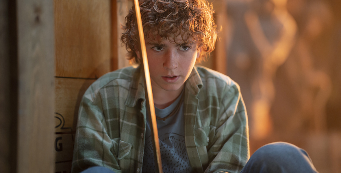 Percy Jackson (Walker Scobell) crouches down before a wooden door with his gold sword drawn. He is looking out intently at something offscreen. He wears a green flannel-print shirt, T-shirt underneath, and jeans.