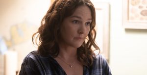 Sally Jackson (Virginia Kull) stands with her body turned toward the right with arms crossed protectively around her waist in a softly lit room. She wears a blue flannel-print long-sleeve shirt over a white T-shirt with a single pearl necklace hanging delicately at her neck.