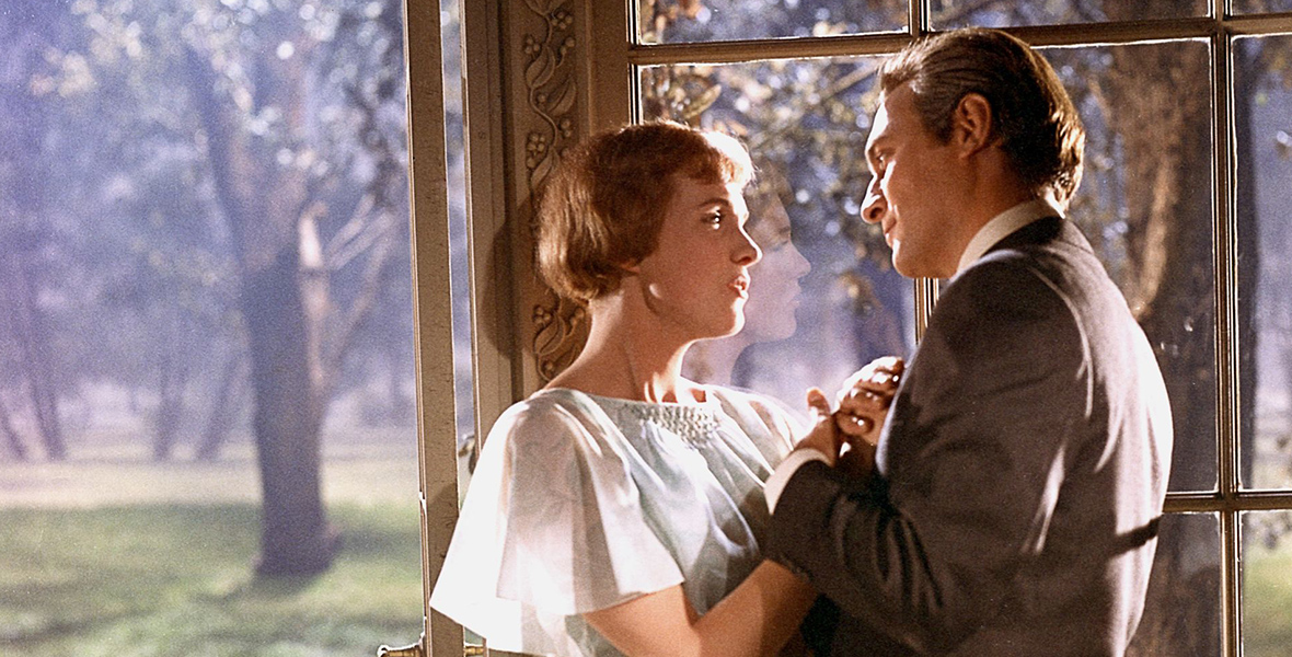 In an image from The Sound of Music, Maria (Disney Legend Julie Andrews) is standing close to Captain Von Trapp (Christopher Plummer) next to a gazebo’s window; a grove of trees can be seen behind them. The pair looks longingly at each other.