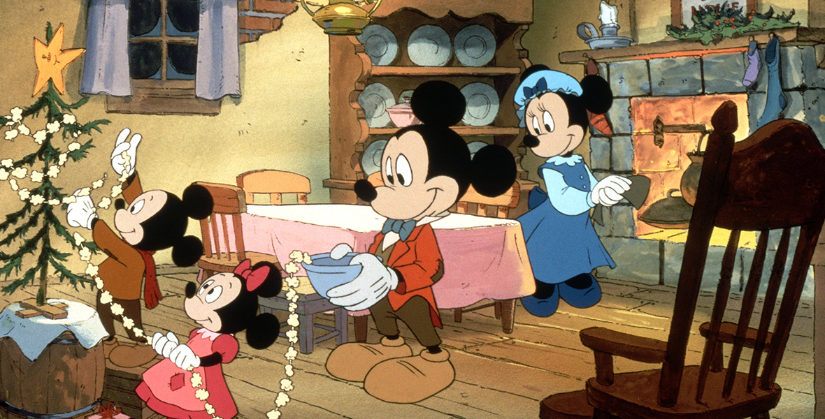 In an image from Mickey’s Christmas Carol, Mickey Mouse as Bob Cratchit and his family are preparing their humble Christmas feast. At left, the children are decorating a small Christmas tree with a string of popcorn, and Minnie Mouse as Mrs. Cratchit is cooking at the hearth.
