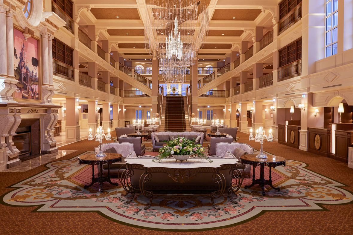 The grand lobby at Disneyland Paris’ Disneyland Hotel features a sparkling chandelier, a stylish table, elegant sofas, and an ornate rug, among other luxurious details.