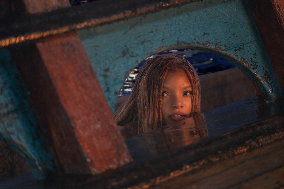 In a scene from The Little Mermaid, Ariel, played by Halle Bailey, peeks her head through an opening on a ship deck and surveys the people onboard.