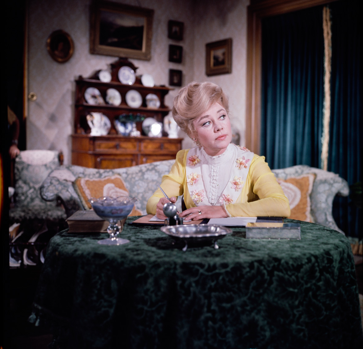 Glynis Johns stars as Winifred Banks in Mary Poppins. She is wearing a yellow dress and is sitting at a table. She is holding a pen in one hand and looking off to the side.