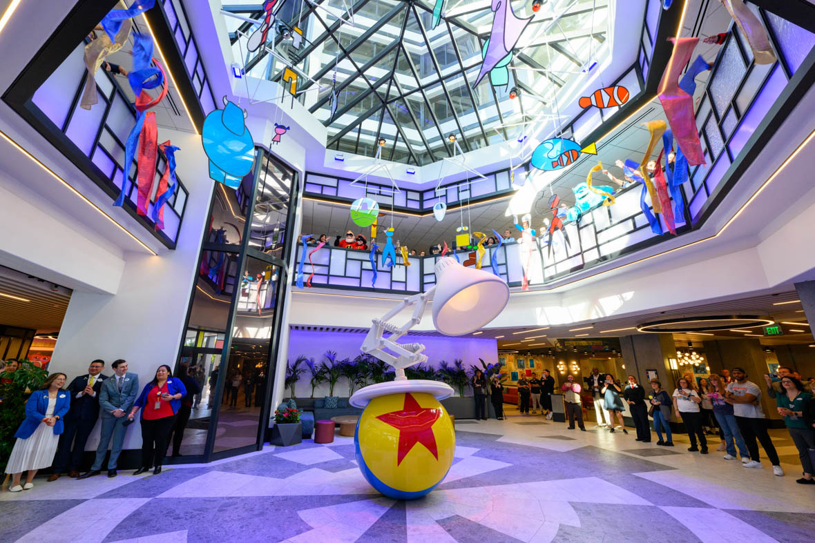 The atrium in the lobby of Pixar Place Hotel is dominated by a sculpture of the white Pixar lamp bouncing atop the iconic yellow Pixar ball, with its blue stripe and red star. Lines of people surround the sculpture and fill the second-floor balcony, where the celebrating figures include characters from the Incredibles movies. Above everything is a domed glass roof.