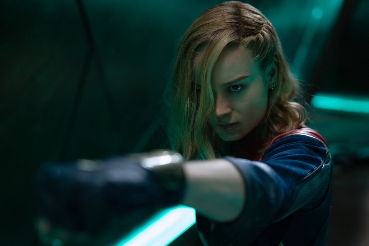 In a scene from The Marvels, Carol Danvers/Captain Marvel, played by Brie Larson, punches with her left arm. Her hair is in her face and she has a steely expression.