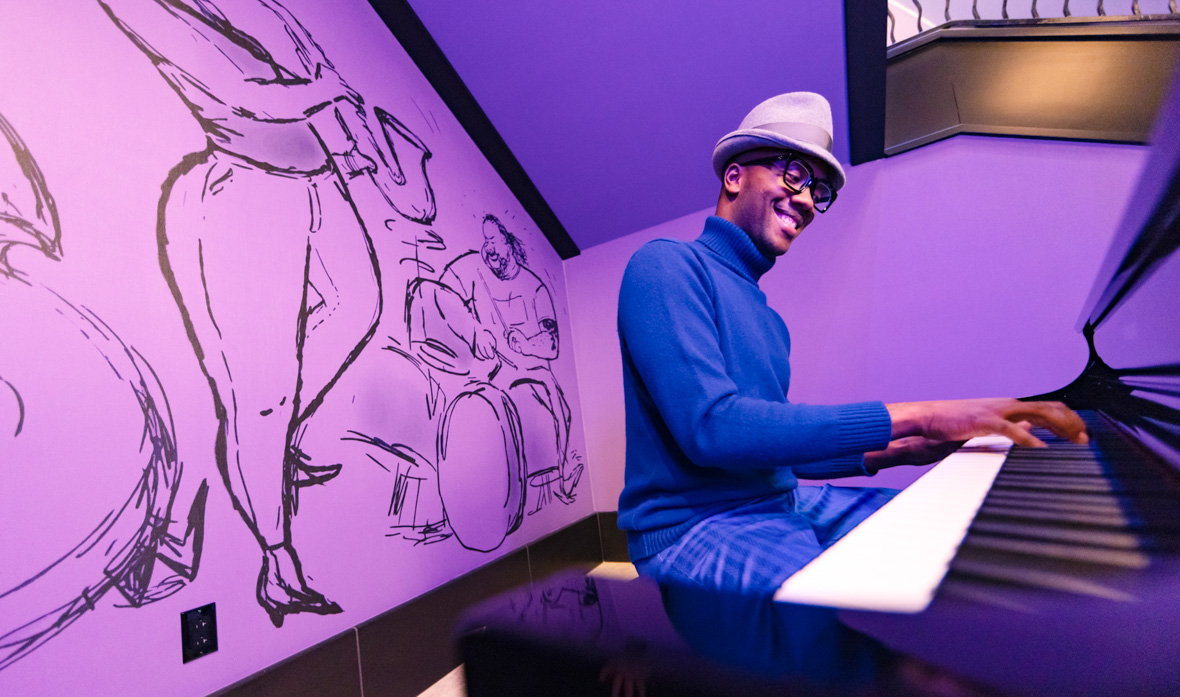 Joe Gardner, wearing a blue turtleneck and matching slacks and his favorite fedora, plays piano in the lobby of Pixar Place Hotel. Behind him is a full-size mural of a jazz trio on a lavender background.