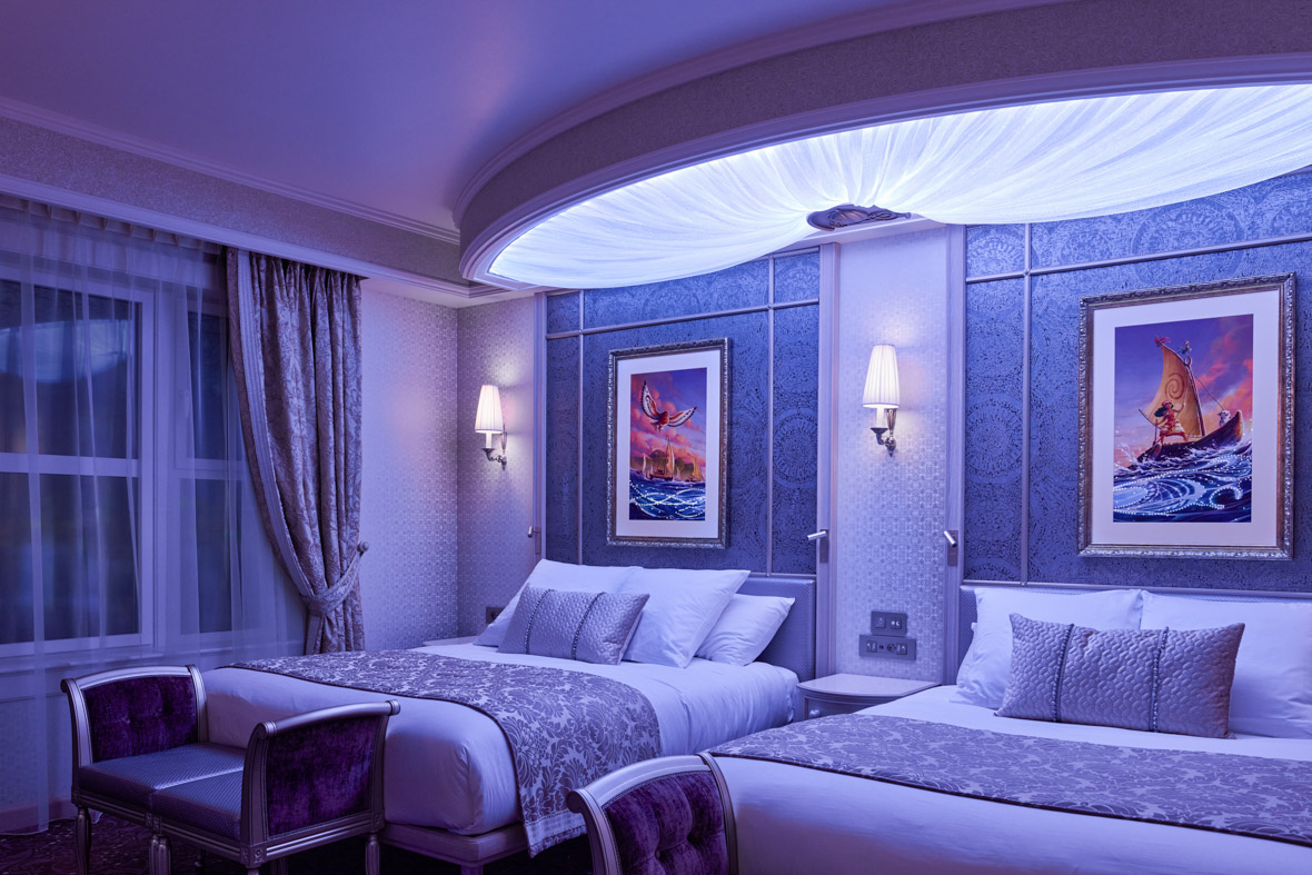 In a Deluxe Room at Disneyland Paris’ Disneyland Hotel, two beds are illuminated by purple light, creating a cozy atmosphere. Framed pictures from Moana hang on the wall.
