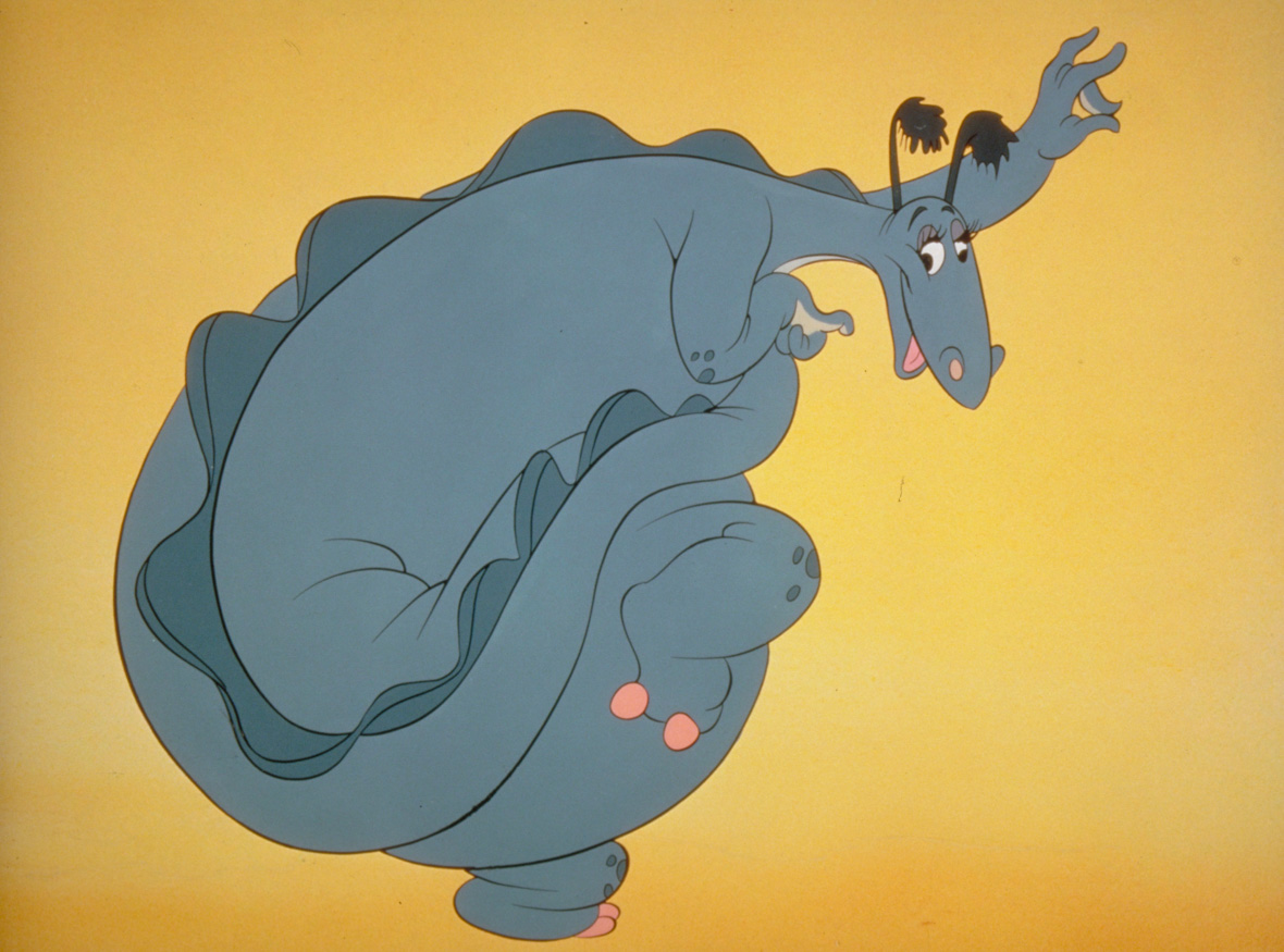 The Reluctant Dragon faces the camera sideways and has one of its legs curled up, as if sitting cross-legged while standing up. The photo’s background is yellow, and The Reluctant Dragon has a warm, happy expression. 