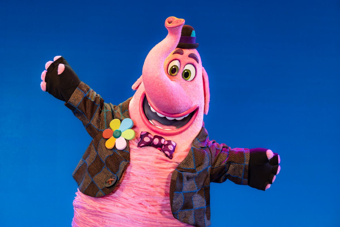 The character Bing Bong from the Pixar Animation Studios film Inside Out stands against a neutral blue backdrop with his arms thrown out. Bing Bong is wearing his usual costume: a patchwork jacket with a big, multicolored daisy pin, fingerless black gloves, bow tie with pink polka dots, and a small Tyrolian hat.