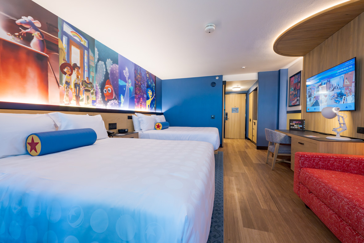 A room at Pixar Place Hotel includes two queen beds, above which is a long mural featuring scenes from Pixar films. On each bed is a cylindrical pillow with the Pixar red star on the end. The beds have white bedspreads and face a flatscreen television and red sofa. At the back of the image is the door to the room.