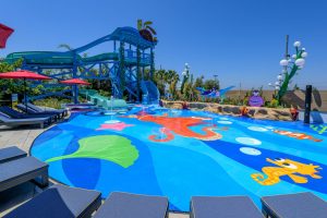 The splash pad at Pixar Place Hotel features characters from Finding Nemo on the blue surface where water sprays out. In the background is Crush’s Surfin’ Slide, a 186-foot-long water slide that curves around and around.