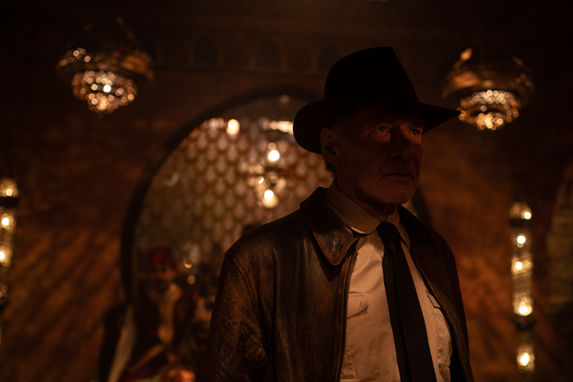 In a scene from Indiana Jones and the Dial of Destiny, Indiana Jones, played by Harrison Ford, wears his signature hat and leather jacket. His face is obscured by shadows.