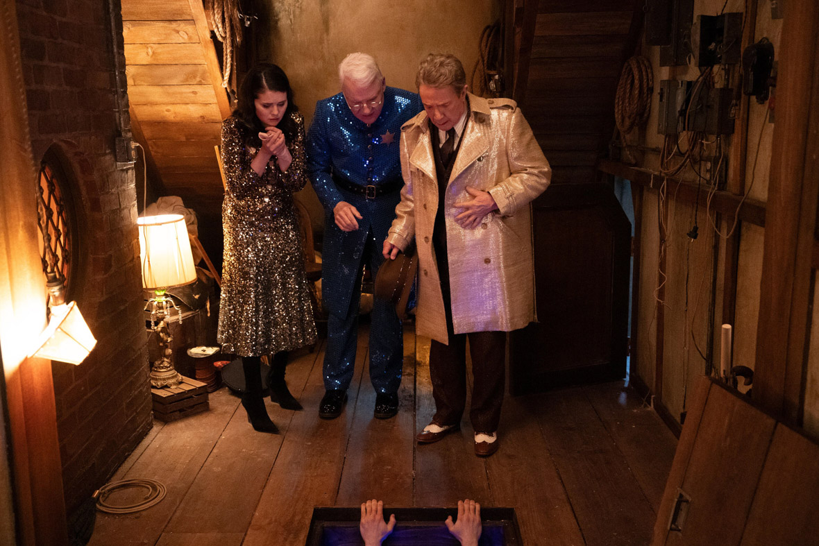 In a scene from Season 3 of Hulu’s Only Murders in the Building, Mabel Mora, played by Selena Gomez, Charles-Haden Savage, played by Disney Legend Steve Martin, and Oliver Putnam, played by Martin Short, stand over a trap door in a backstage area. A person’s hands are reaching up through the trap door and holding onto the wooden floor.