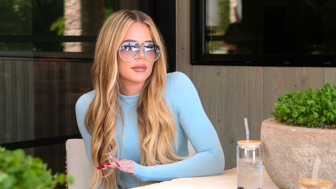 In a scene from Hulu's The Kardashians, Khloé Kardashian wears a blue, long-sleeved top and blue aviator sunglasses. A glass of water is on the table in front of her.