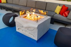 On the pool deck at Pixar Place Hotel is one of four character-themed firepits, each one a cube of what appears to be concrete with the face of a Pixar character on one side. The firepit depicted has the face of Anger from Inside Out, and a gas fire is burning atop his head. Beside the firepit are two black stools and behind it is a gray banquette.