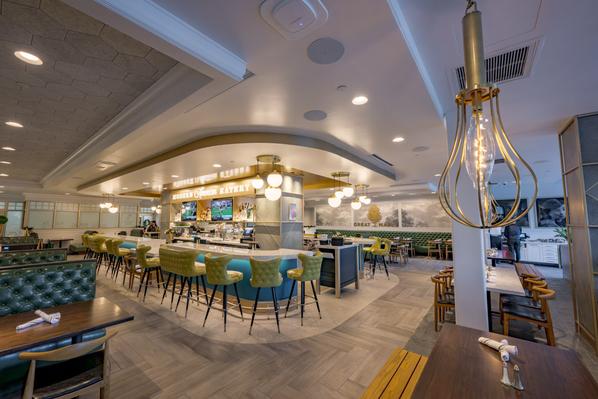 The Great Maple restaurant at Pixar Place Hotel includes a four-sided bar with blue-backed bar stools. Banquettes line the side of the room. In the foreground is a brown table with a wire-frame light fixture hanging above it.