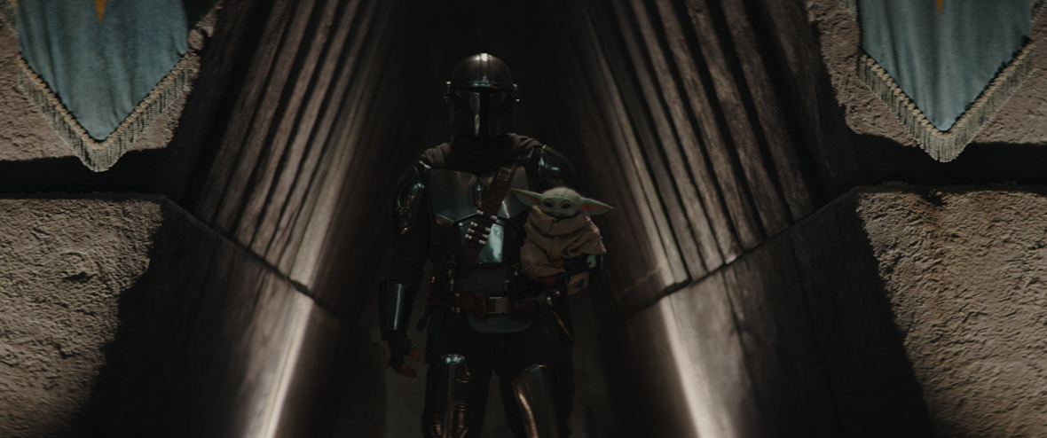In a scene from Season 3 of Star Wars: The Mandalorian, Din Djarin, played by Pedro Pascal, wears full Mandalorian armor. He is holding Grogu, a small green creature.