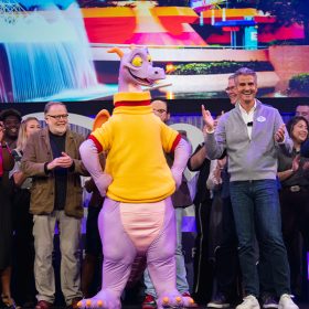 Dave Goelz, Josh D’Amaro, and a crowd of Disney Parks and Experiences cast members stand on stage surrounding Figment the dragon. Behind them all, an image of EPCOT’s Imagination Pavilion is projected on a screen.