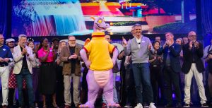 Dave Goelz, Josh D’Amaro, and a crowd of Disney Parks and Experiences cast members stand on stage surrounding Figment the dragon. Behind them all, an image of EPCOT’s Imagination Pavilion is projected on a screen.