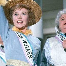 Glynis Johns stars as Winifred Banks in Mary Poppins. She wears a straw bonnet and a white sash that reads "Votes for Women," trimmed in pink and blue ribbon. Her right arm is outstretched upward as she bursts into song. Winifred is flanks by two housekeepers.