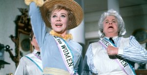 Glynis Johns stars as Winifred Banks in Mary Poppins. She wears a straw bonnet and a white sash that reads "Votes for Women," trimmed in pink and blue ribbon. Her right arm is outstretched upward as she bursts into song. Winifred is flanks by two housekeepers.