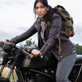 In an image from Marvel Studios’ Echo, Maya Lopez (Alaqua Cox) is on the back of a motorcycle, looking at the camera. She’s wearing a blue jacket, a plaid shirt, and jeans, and has an army green backpack on her back.