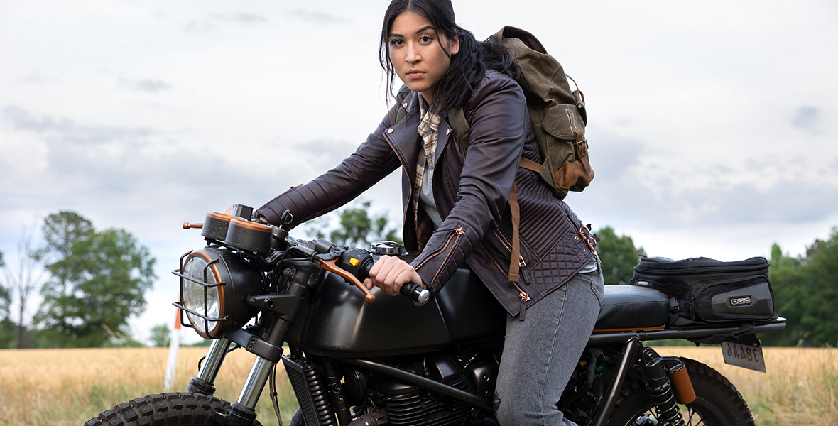 In an image from Marvel Studios’ Echo, Maya Lopez (Alaqua Cox) is on the back of a motorcycle, looking at the camera. She’s wearing a blue jacket, a plaid shirt, and jeans, and has an army green backpack on her back.