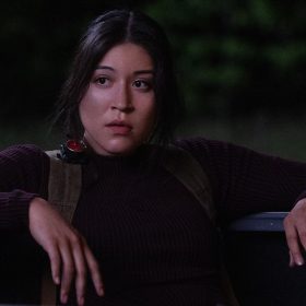 In an image from Marvel Studios’ Echo, Maya Lopez (Alaqua Cox) is leaning back on what appears to be the bed of a truck, wearing dark clothing and looking at something off camera to the left.