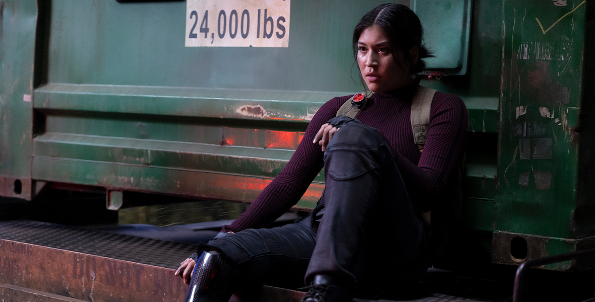 In an image from Marvel Studios’ Echo, Maya Lopez (Alaqua Cox) is leaning back against what appears to be a truck—there’s a sign on the back of the truck stating “Forklift Floor Rating: 24,000 pounds.” She is wearing dark clothing, with one foot pulled up on the ledge she’s sitting on.