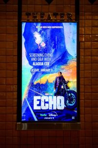 A poster outside the theater at the Autry Museum of the American West, announces the screening event and Q&A with Echo’s lead actress Alaqua Cox showcasing Echo, dressed in leather jacket and dark pants, her hair pulled back astride a motorcycle at the edge of railroad tracks. A vibrant sunset skey and the outline of Kingpin are in the foreground.