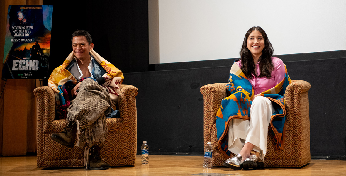 (From left to right) Chaske Spencer (Henry) and Alaqua Cox (Echo) sit on stage in plush armchairs facing the audience and smiling during a Q&A panel for the Marvel Studios series  Echo at the Autry Museum of the American West. Both actors are draped in tribal blankets.