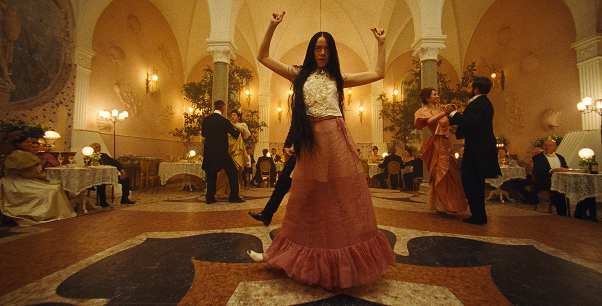 In a scene from Poor Things, a raven-haired Bella Baxter, played by Emma Stone, raises her arms and snaps her fingers while standing in the center of the dance floor. Two couples dance behind Bella as several other people watch from surrounding dining tables.