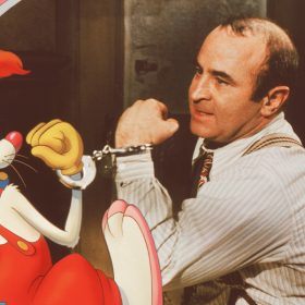 In an image from Disney’s Who Framed Roger Rabbit, Roger Rabbit (voiced by Charles Fleischer), a white cartoon rabbit, and Eddie Valiant (Bob Hoskins), a human man, stare at each other, both holding up their arms to show that they are handcuffed together.