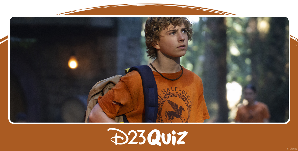 Percy Jackson stands in front of a cabin at Camp Half-Blood, wearing his backpack on one shoulder with a contemplative expression on his face. He is wearing an orange Camp Half-Blood T-shirt, as are the out-of-focus campers in the background of the image.