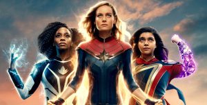 In a promotional poster for Marvel Studios' The Marvels, Monica Rambeau, played by Teyonah Parris, Carol Danvers, played by Brie Larson, and Kamala Khan, played by Iman Vellani, strike classic Super Hero poses. They respectively emit white, gold, and purple light.