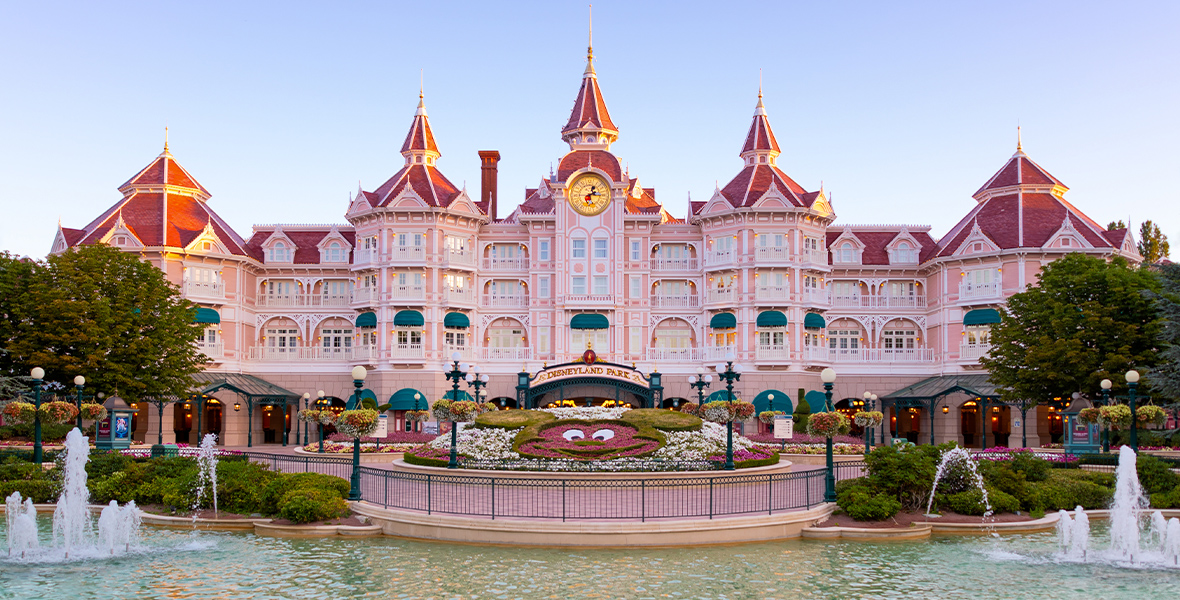 The reimagined Disneyland Hotel at Disneyland Paris overlooks a lake. The majestic pink building features a Mickey Mouse clock, showcasing architectural grandeur.