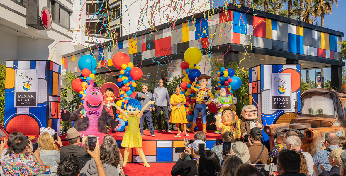 On a stage set up in front of Pixar Place Hotel at Disneyland Resort, a host of Pixar characters raise their arms as streamers fly through the air in celebration of the hotel’s grand opening. The characters include Bing Bong and Joy from Inside Out, Woody and Buzz from the Toy Story movies, and Russell and Carl from Up.