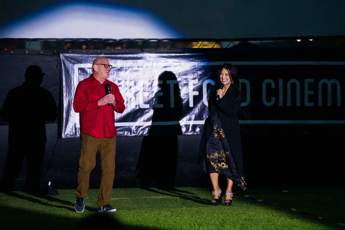 Head of D23, Michael Vargo, and Disney Legend Ming-Na Wen stand in front of an outdoor movie screen and a banner for “Street Food Cinema.” Both are smiling and holding microphones. 