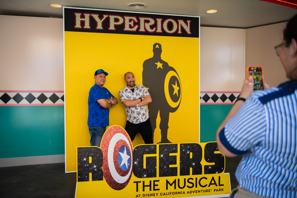 Two men pose in front of a photo backdrop designed to look like a playbill for Rogers: The Musical. A cast member is out of focus in the right side of the photo, holding up a camera phone to take the men’s photo. 