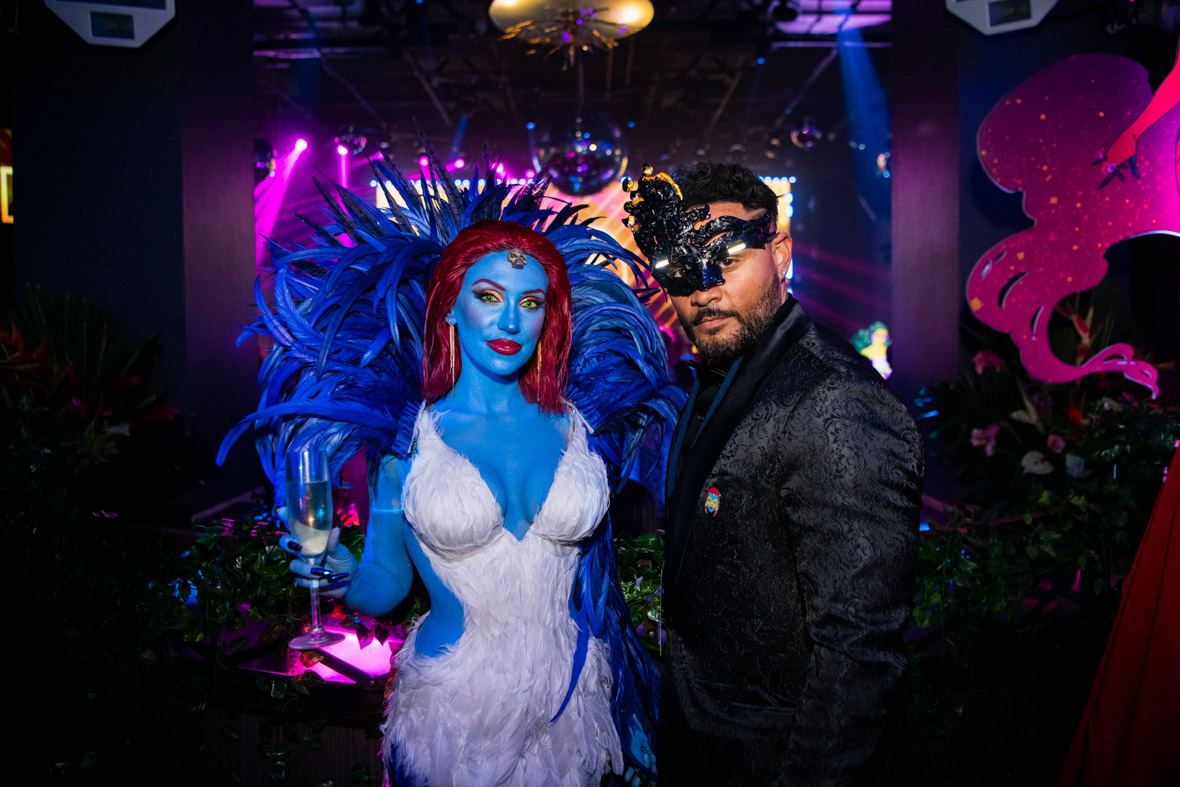 A woman dressed as the X-Men character Mystique holds champagne glass and poses with a man in a black suit and mask. They both stand in front of a colorful dance floor with a disco ball above it. 