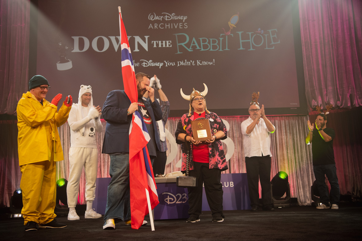 D23 and Archives team members, dressed like various characters from the EPCOT attraction Malestrom, stand around Kevin Kern and Becky Cline from the Walt Disney Archives. Kevin is holding the Norway flag and laughing, while Becky is wearing Viking horns and holding a plaque honoring Kevin’s years of service. 