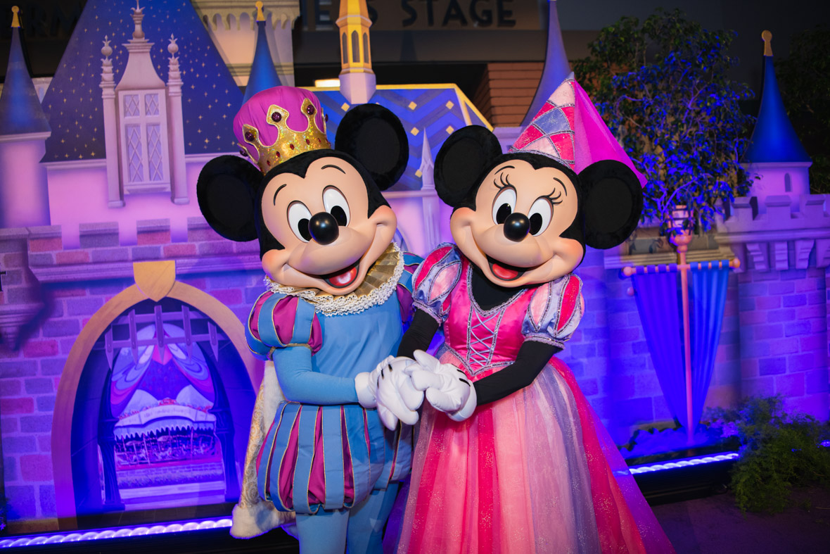 Mickey and Minnie, dressed like a prince and princess, pose in front of a photo backdrop depicting Sleeping Beauty Castle. 