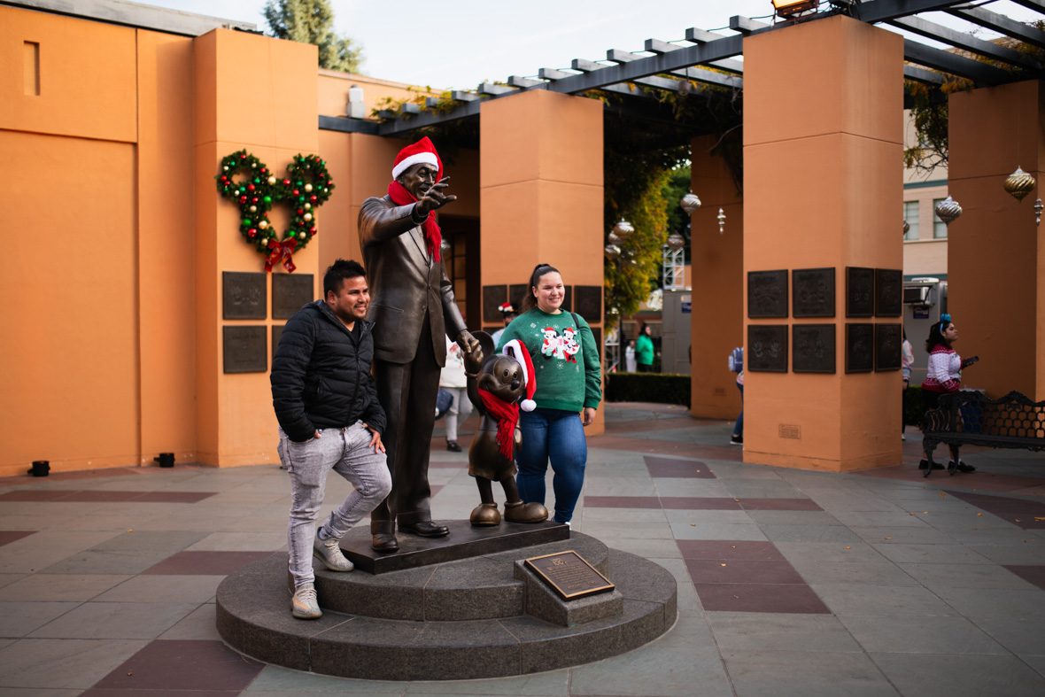 Two guests pose with the “Partners” statue in Legends Plaza at the Walt Disney Studios lot. The statue is decorated for Christmas, with both Walt and Mickey wearing Santa hats and scarves. 