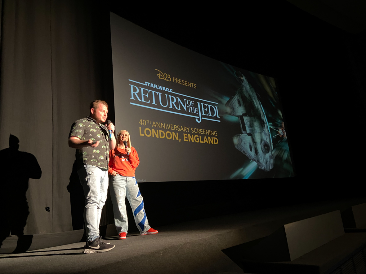 D23’s Justin Arthur stands on stage with Ashley Eckstein. Both hold microphones and stand next to a screen depicting an image of the Millennium Falcon and the text “D23 Presents Star Wars: Return of the Jedi 40th anniversary screening in London, England.”