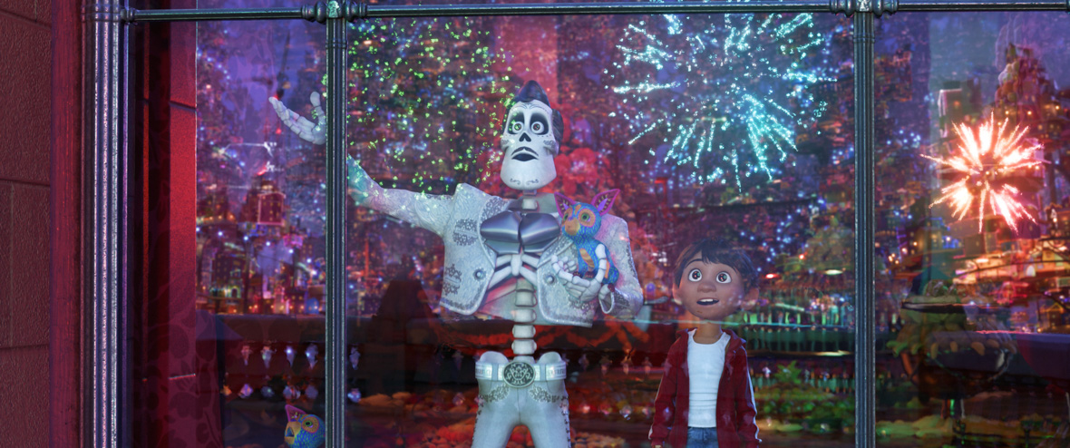 In an image from Disney and Pixar’s Coco, Ernesto de la Cruz (voiced by Benjamin Bratt), a skeleton in a white jacket and white pants, and Miguel (voiced by Anthony Gonzalez), a human boy in a red hoodie, stare out a window with awe. The colorful fireworks they are watching are reflected in the glass of the window.