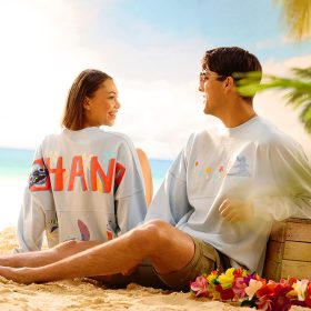 A young woman and man sit on a sandy beach surrounded by palm trees and tropical flowers, both smiling and wearing matching Stitch-themed Disney Spirit Jerseys. The woman’s shirt reads “’OHANA” across the back with colorful tropical designs, while the man’s shirt features Stitch surfing. A wooden crate sits beside them with the ocean and sky in the background.