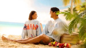 A young woman and man sit on a sandy beach surrounded by palm trees and tropical flowers, both smiling and wearing matching Stitch-themed Disney Spirit Jerseys. The woman’s shirt reads “’OHANA” across the back with colorful tropical designs, while the man’s shirt features Stitch surfing. A wooden crate sits beside them with the ocean and sky in the background.