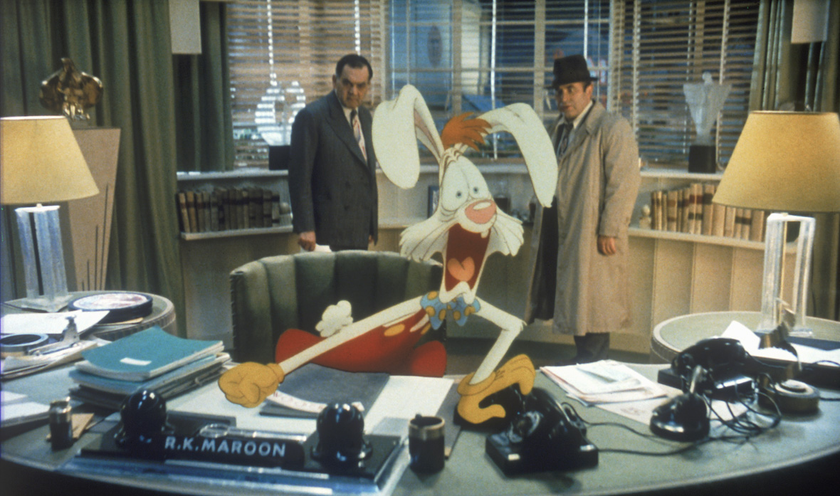 In an image from Disney’s Who Framed Roger Rabbit, Roger Rabbit (voiced by Charles Fleischer), a cartoon white rabbit, sits at the live-action desk of R.K. Maroon (Alan Tilvern). Roger’s expression is frozen in mid-scream, while humans R.K. Marron and Eddie Valiant (Bob Hoskins) stand by the window behind Roger, both staring in shock at him.